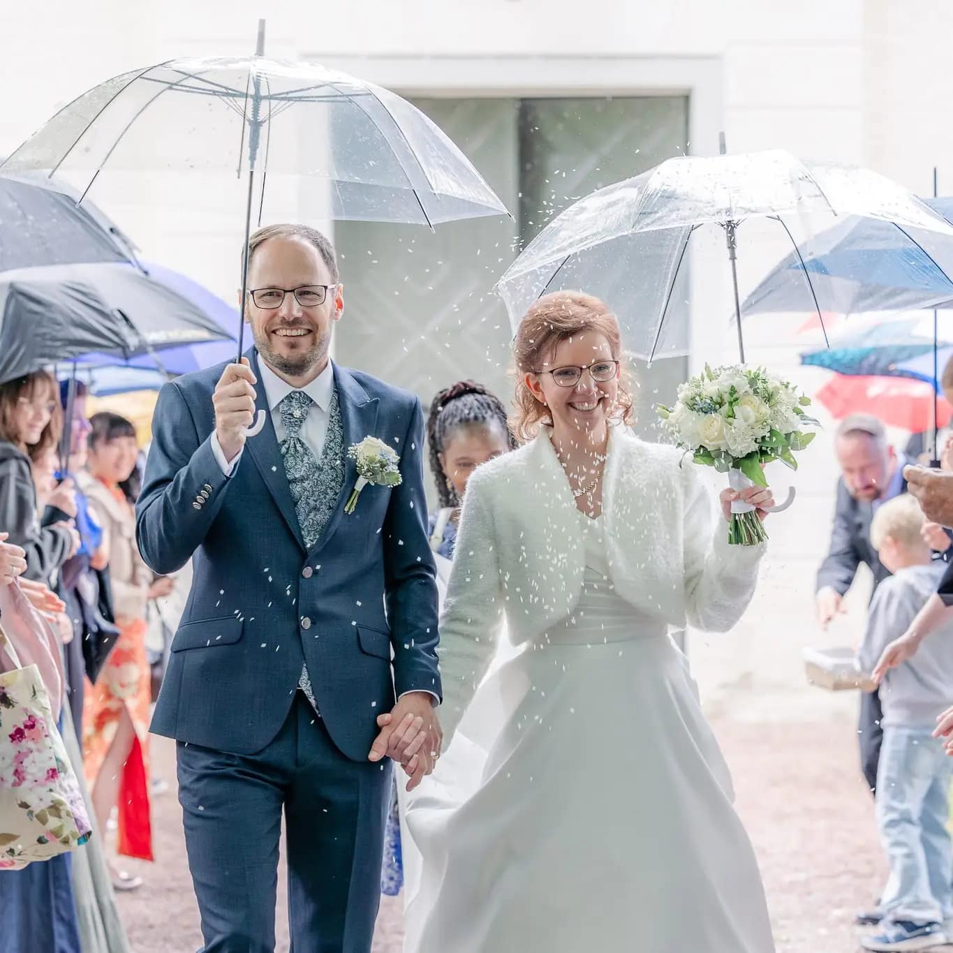 Brudpar kommer ut ur kyrka med transparenta paraplyer medans gästerna kastar ris